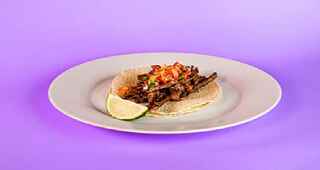 A taco with tomatoes on top with a slice of lime by it on a plate with a purple background.