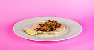 A taco with sweet potato shaving on top with a slice of lime on the side on a pink background.